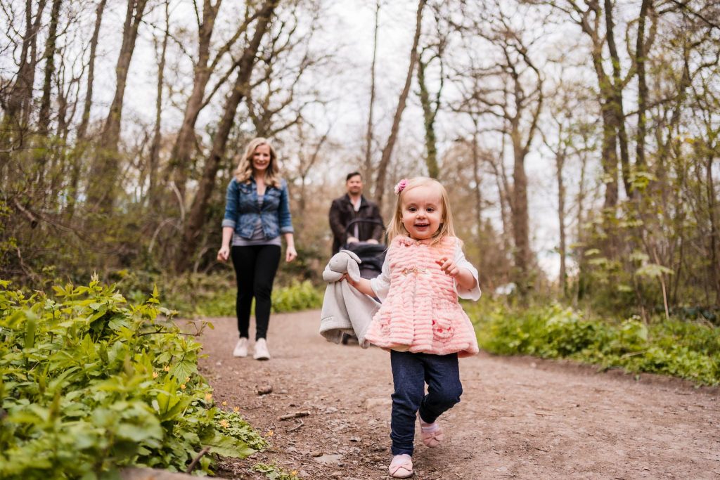 A young girl in a pink coat joyfully runs along a forest path, clutching a stuffed animal. Behind her, two adults casually stroll, enveloped by lush trees and a serene woodland atmosphere—a perfect moment captured by Leicester family photographer.