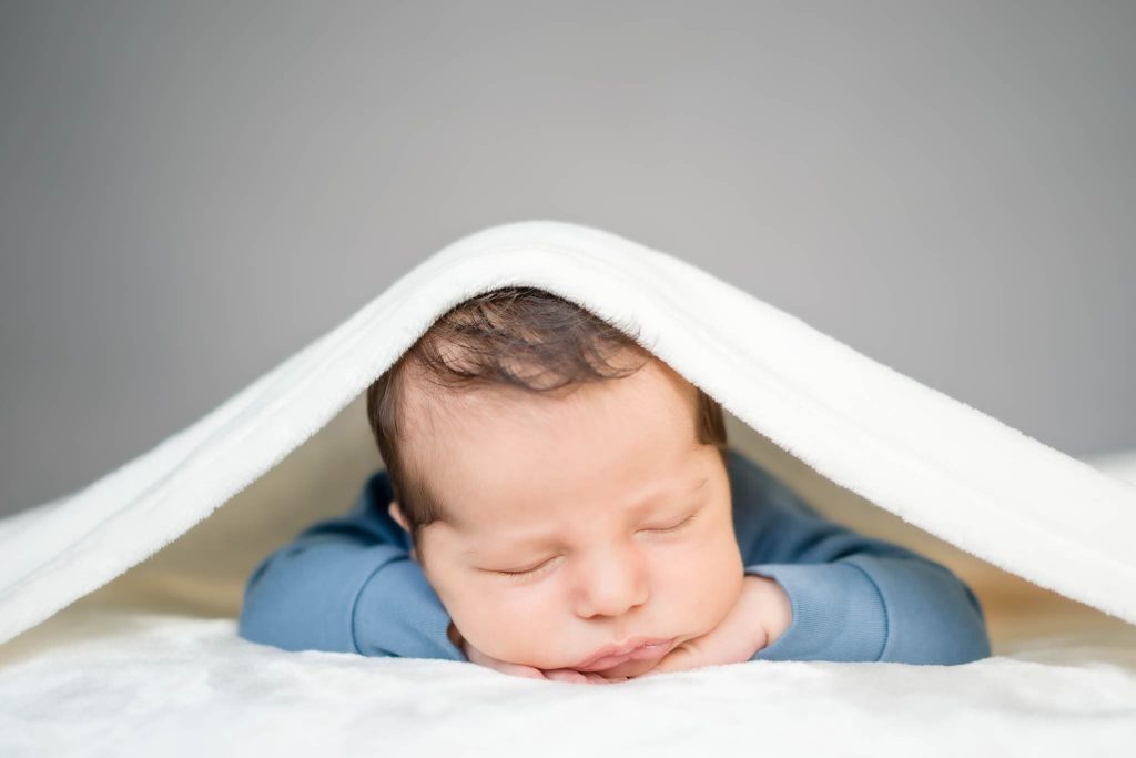 A sleeping baby in a blue outfit is nestled under a soft white blanket, their head resting on tiny hands, while the blanket forms a cozy canopy. Captured by a Leicester family photographer, the softly blurred neutral background enhances the serenity of this precious moment.