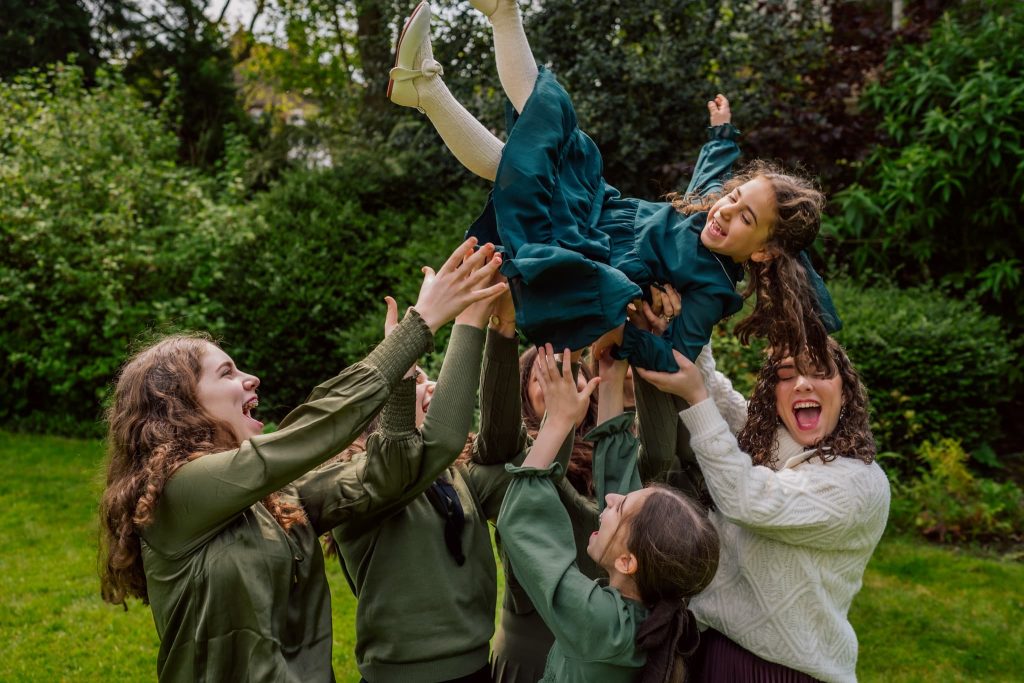 In a lush garden,a group of smiling people playfully lift a young girl in a green dress, captured perfectly by a Leicester family photographer. Surrounded by trees and greenery, the atmosphere is joyful and celebratory.