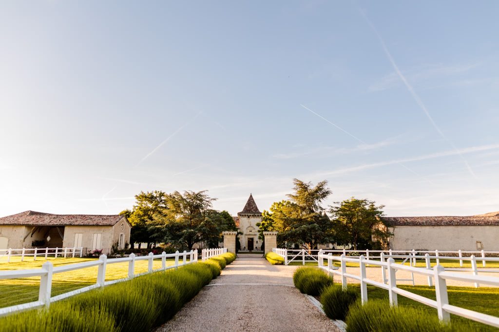 The picturesque Chateau Soulac, France, with a central tower stands at the end of a gravel path lined by manicured hedges and white fences. Surrounded by green lawns and trees, the scene is bathed in warm sunlight under a clear blue sky—a perfect backdrop for a destination wedding photographer.