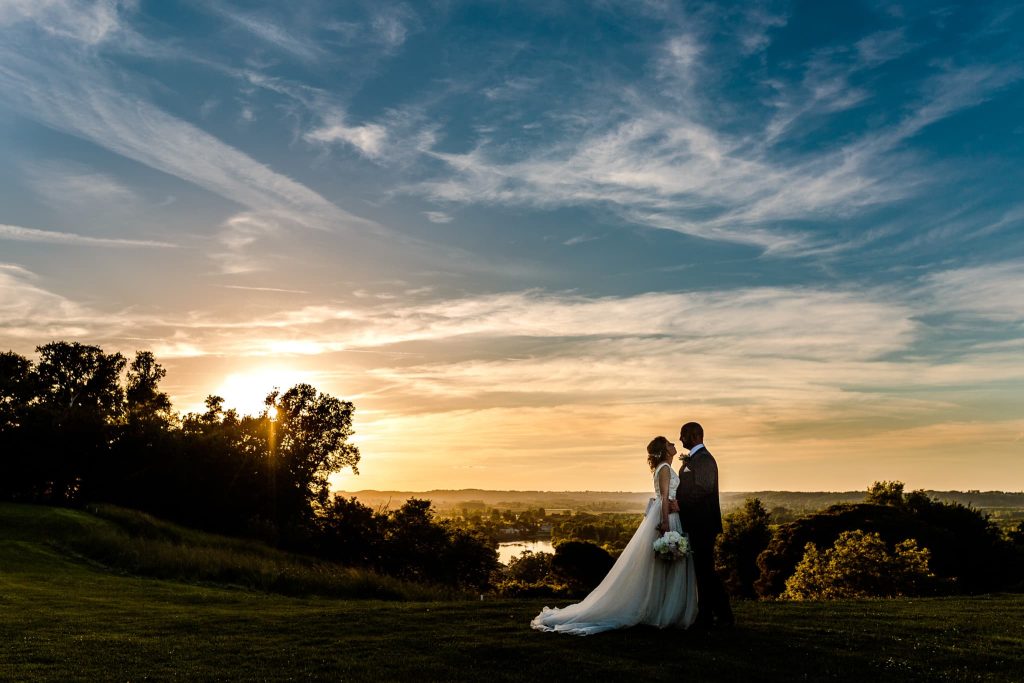 A bride and groom stand closely on a grassy hill at sunset, the perfect scene for a France destination wedding photographer. The sun casts a warm glow over trees and a river in the background, with wispy clouds painting the sky.