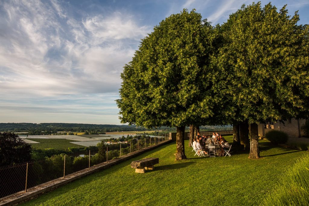 A group of people sits at a table under large, leafy trees on a grassy hillside overlooking a landscape of vineyards and fields—an idyllic scene perfect for a France destination wedding. The sky is partly cloudy, and there is a fence running along the edge of the grassy area.