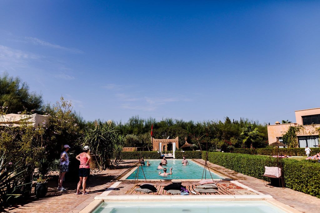 A group of people relax in a rectangular outdoor pool surrounded by greenery and bushes under a clear blue sky, the perfect scene for a Morocco destination wedding. Two people stand talking on the poolside. A small structure is visible in the background, with pillows and towels lining the pools edge.