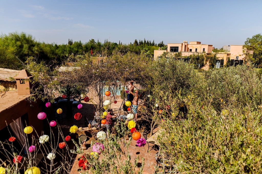 A vibrant outdoor setting with colorful lanterns and flowers hanging from trees. People gather in a courtyard surrounded by greenery and Mediterranean-style buildings under the clear blue sky, perfect for a Marrakesh destination wedding photographer to capture unforgettable moments.