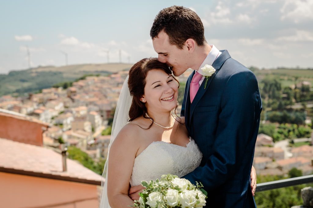 A bride and groom share a joyful embrace on their wedding day. The groom kisses the brides forehead as she holds a bouquet of white flowers. An Italy destination wedding, set against a scenic hillside town and blue sky, is beautifully captured by the photographer.