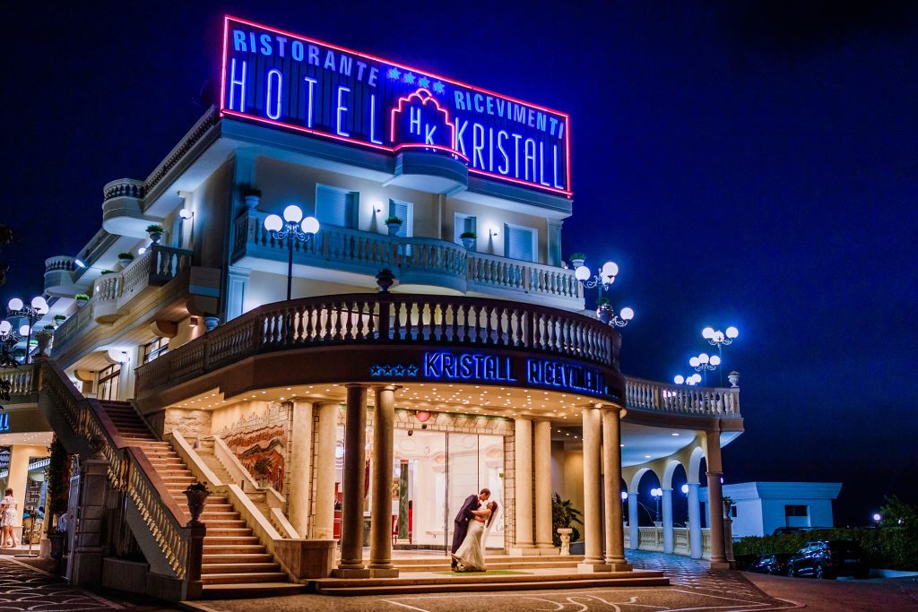 A brightly lit hotel with a neon sign reading Ristorante Ricevimenti Hotel Kristall at night. Perfect for an unforgettable European destination wedding, the building is adorned with glowing lights. A couple stands in an embrace under the entrance, captured beautifully by their photographer.