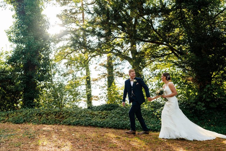 Bride and groom walk hand in hand through a sunlit forest at their Whatton House Wedding. The bride, in a white gown and veil, holds a bouquet while the groom dons a dark suit with a boutonniere. Sunlight filters through the trees, casting a warm glow on this enchanting scene.